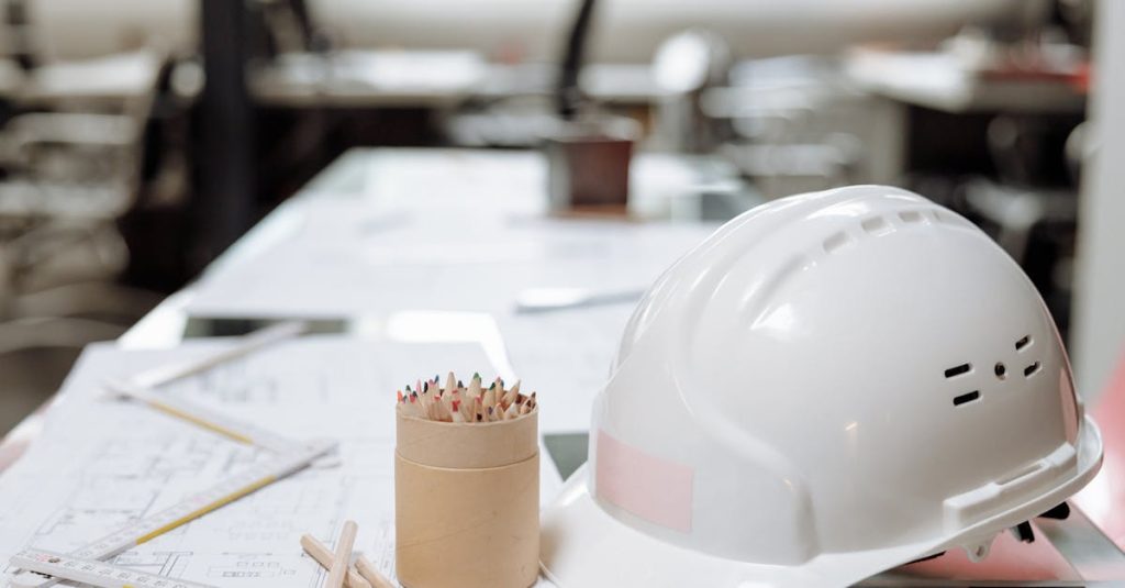 Close-up of an office desk with a white helmet, colored pencils, and architectural plans.